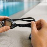 Hands adjusting the split strap and buckle of swim goggles beside a pool